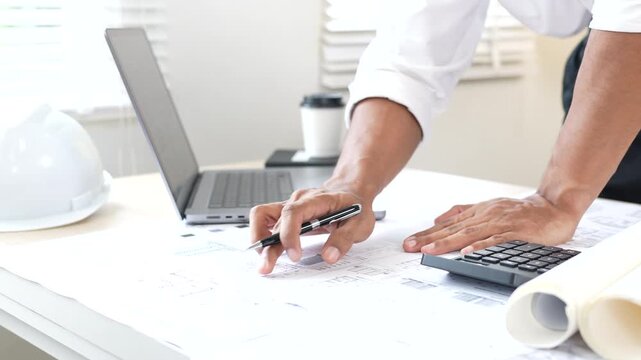 Architect reviewing construction plans at a modern office desk with blueprints, laptop, and engineering tools for a building project.