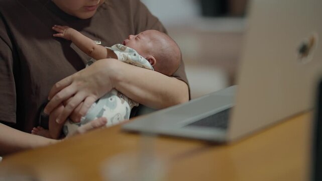 Mother holds newborn while working on laptop. Baby sleeps peacefully in her arms. Soft lighting enhances quiet, caring moment. Ideal for parenting, new mother, or work-life balance scenes