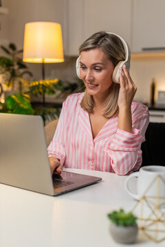 Adult woman at night home works in design tool on laptop with headphones moves layers tweaks layout smiles. Caucasian girl at table on sofa at evening nods music applies edits saves version progress.