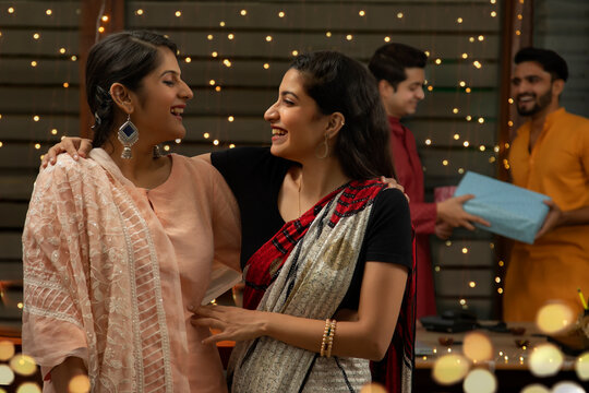 YOUNG WOMEN LOOKING AT EACH OTHER AND LAUGHING WITH MALE COLLEGUES GIFTING EACH OTHER BEHIND