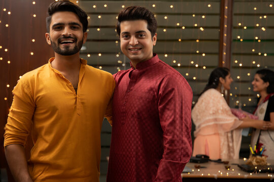 TWO MALE COLLEGUES LOOKING AT CAMERA AND POSING WITH FEMALE COLLEGUES GIFTING EACH OTHER IN THE BACKGROUND