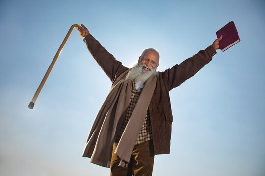 A BEARDED OLD MAN LOOKING AT CAMERA WHILE HAPPILY RAISING HANDS