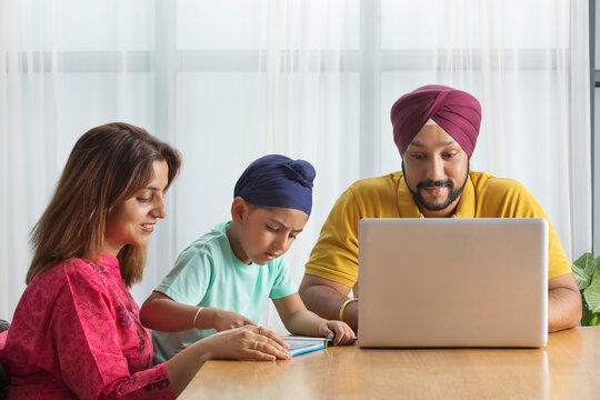 A SIKH BOY READING WITH MOTHER WHILE FATHER IS WORKING ON HIS LAPTOP