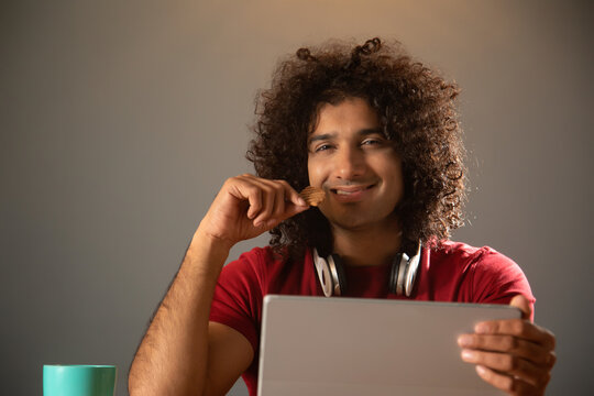 A CURLY HAIRED MAN HAPPILY EATING CHIPS WHILE HOLDING DIGITAL TABLET