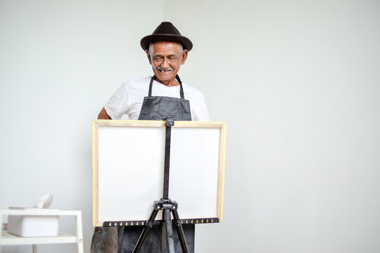 Smiling Elderly Asian Man Wearing Hat and Apron Standing Behind Blank White Canvas on an Easel
