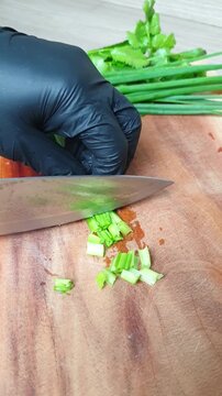 Chef wearing black gloves finely chopping fresh culantro herbs on a wooden cutting board with a sharp knife in the kitchen