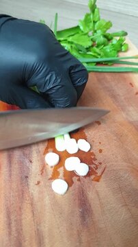 close up view of a hand in black nitrile gloves slicing spring onions on a wooden cutting board with a knife