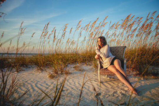 Young woman in shorts and a jumper sitting on a chair on a sandy beach amongst the long grass, Florida, USA