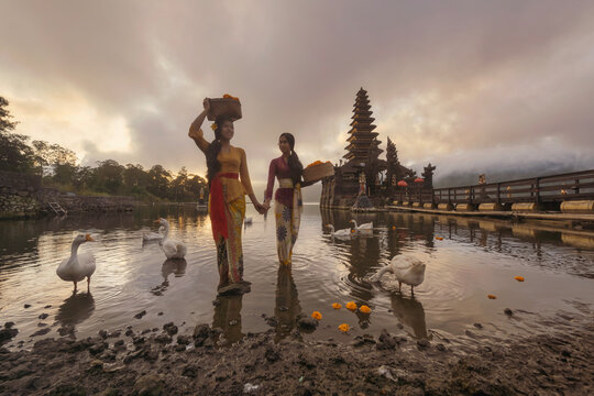 Two smiling Balinese women in a traditional kebaya batik dress standing in front of a temple in shallow water carrying baskets of flowers, Indonesia