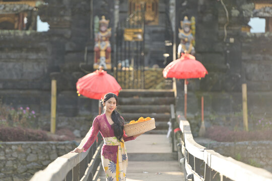 Portrait of a smiling Balinese woman in a traditional kebaya batik dress standing in front of a temple holding a basket of freshly picked flowers, Indonesia