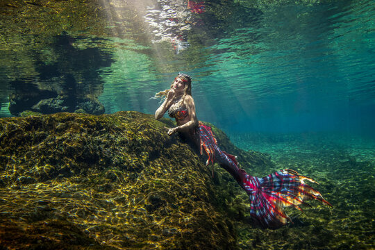 Side view of a beautiful mid adult woman with a mermaid tail swimming along the ocean floor