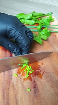 Chef wearing black gloves finely chopping fresh cilantro leaves on a wooden cutting board with a sharp knife in the kitchen