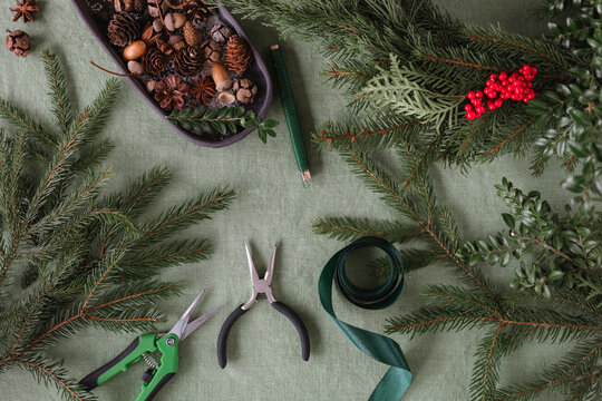 Overhead view of various items on a table for making a Christmas wreath including assorted foliage, pinecones, ribbon, ribbon, pruning shears and pliers
