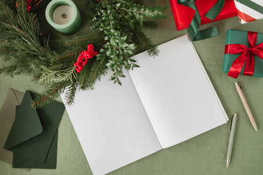 Overhead view of an open blank notebook on a table with a candle in a Christmas wreath, wrapped Christmas gifts and assorted stationery