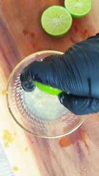 A hand in a black glove squeezing fresh lime juice into a clear glass bowl on a wooden cutting board