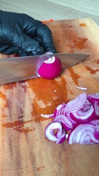 A chef wearing a black glove slicing a fresh red onion on a wooden cutting board with a sharp knife