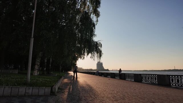 Man walks along a paved river embankment path away from camera at sunset. Sun rays filtering through trees, long shadows and a cast iron railing
