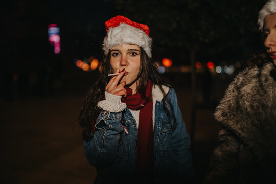 A young woman wearing a Santa hat smokes a cigarette while standing outdoors at night. She pauses during a festive evening with friends in a dimly lit street scene.