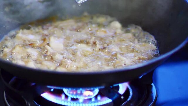 Pieces of food sizzling and deep frying in hot oil in a black wok on a gas stove. The oil is bubbling vigorously, creating a dynamic and appetizing scene