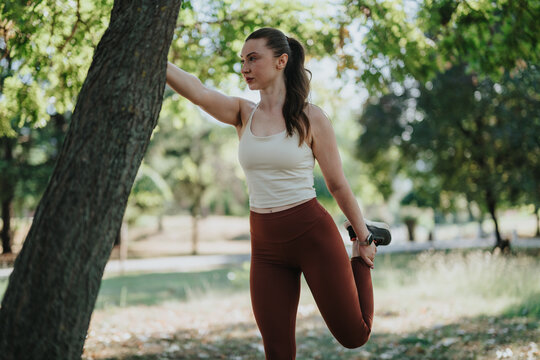 A young woman performs a standing quad stretch while holding her foot in a leafy park. She stretches one leg for an outdoor fitness and sport training session.