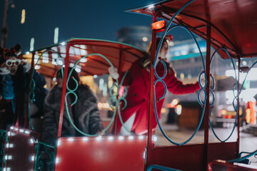 Fototapeta premium Two girls ride a small decorative tram at a Christmas market at night. The festive red carriage glows with lights as they enjoy the holiday attraction.