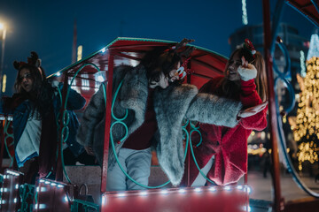 Fototapeta premium Three girls wearing festive winter clothes ride a brightly lit amusement train at night. They laugh and lean out of the carriage amid holiday lights and decorations.