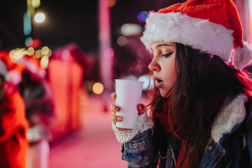 Fototapeta premium A young girl wearing a Santa hat holds a steaming paper cup while standing outdoors at night. A festive holiday market scene with Christmas lights and bokeh in the background.