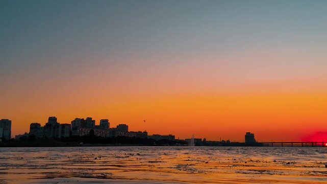Wide low-angle panning shot from high-rise city skyline across the water to a vibrant red sunset setting over a long river bridge.