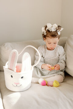 Toddler girl opens colourful Easter eggs beside a bunny basket at home