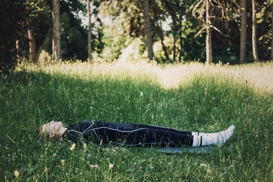 Woman in athletic wear lies on mat among tall grass in park. Yoga nidra restoration, deep relaxation practice, sleep meditation outdoor.
