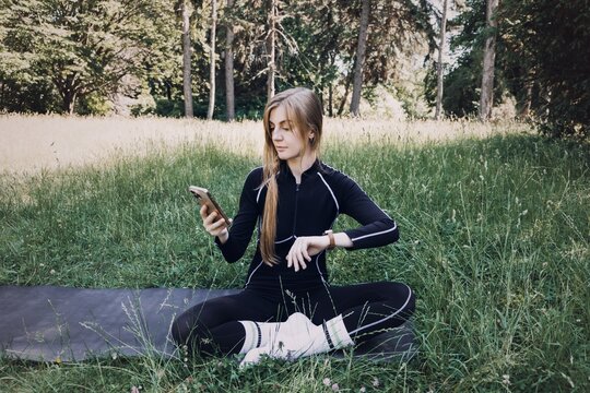 Woman sits on yoga mat in grass looking at smartwatch and holding smartphone. FemTech app tracking, menstrual cycle app, women health technology, women wellness tracker.