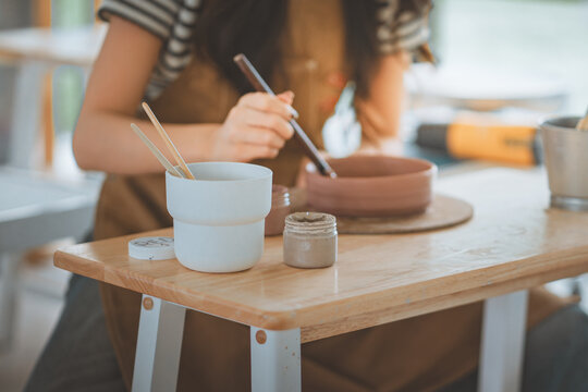Close up of Asian woman hands painting handmade ceramic clay bowl with paintbrush in pottery workshop studio class, young female artist creating handcrafted stoneware dish for art therapy.