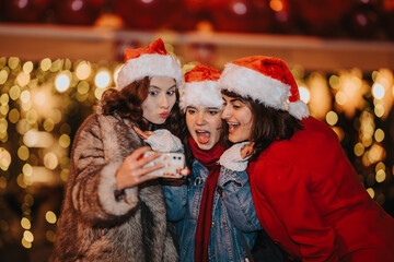 Fototapeta premium Three girls wearing Santa hats take a cheerful selfie together. The friends pose and laugh in front of warm Christmas lights at a holiday market.