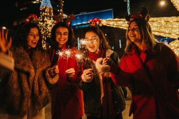 Fototapeta premium Girls hold lit sparklers and laugh while wearing festive headbands and winter coats. A joyful group of friends celebrates outdoors at night surrounded by holiday lights.