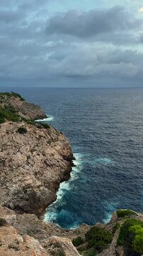 Morning light illuminates Capdepera Lighthouse on rocky coast of Mallorca. Vertical travel scene dark blue sea dramatic gray sky and Mediterranean atmosphere.