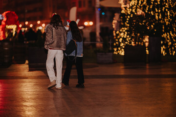 Fototapeta premium Two girls walk together at night next to illuminated holiday decorations. They stroll through a city square with warm festive lights in the background.