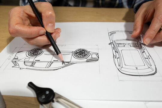 Hands of engineer sketching automotive vehicle prototype with pencil on paper at desk. Industrial designer developing new car blueprint drafts near measuring tools and rulers.