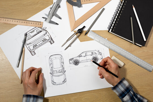 Hands of engineer sketching automotive vehicle prototype with pencil on paper at desk. Industrial designer developing new car blueprint drafts near measuring tools and rulers.