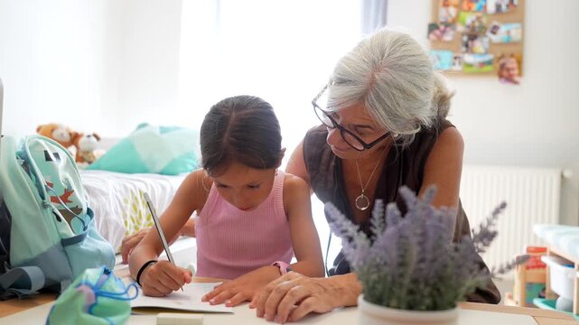 Loving grandmother with grey hair and glasses assisting her young granddaughter with her school assignments at a desk in her bedroom, fostering a positive learning environment