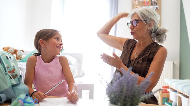 Caring grandmother assisting her happy granddaughter with schoolwork after school, sitting at a desk in the child's bedroom, celebrating success with a high five and expressing joy together