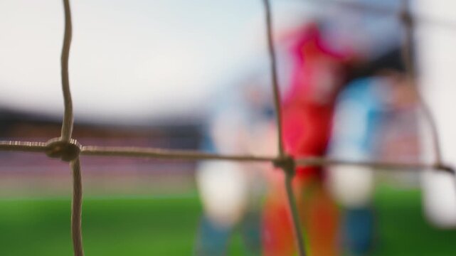Selective focus of soccer ball crossing line of goal frame on pitch, goal shot hitting net, red soccer players of winning team cheering near defeated opponents, fallen goalie in blurred motion.