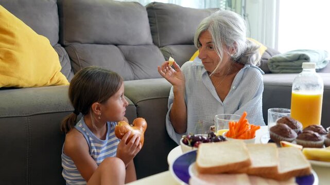 Loving grandmother and her granddaughter enjoying a snack together in the living room. Adorable intergenerational scene capturing a moment of conversation and affection while sharing a healthy meal