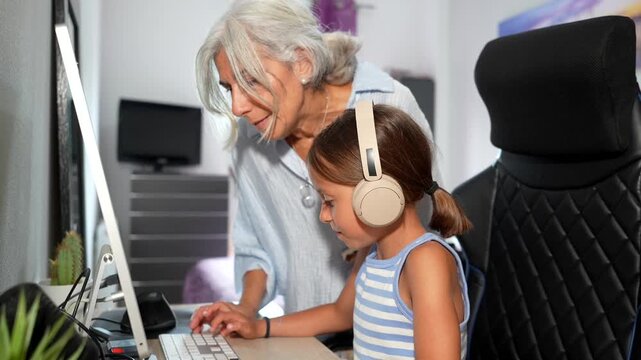 Elderly woman helping her young granddaughter with homework on a desktop computer at home, fostering a positive intergenerational bond through shared learning and modern technology