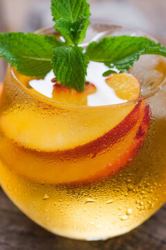 A glass filled with a beverage made of peach slices and mint leaves sits on a wooden table. The drink is cold and has condensation on the outside of the glass.