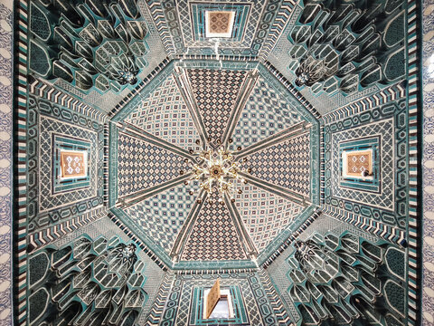 Ornate tiled ceiling inside a mausoleum at Shah-i-Zinda necropolis in Samarkand, Uzbekistan. Intricate geometric mosaic patterns and golden chandelier viewed from below. Ideal for travel.
