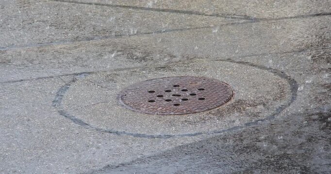 Rain drops fall on a round rusty metal sewer manhole cover on asphalt street during heavy downpour storm outdoors