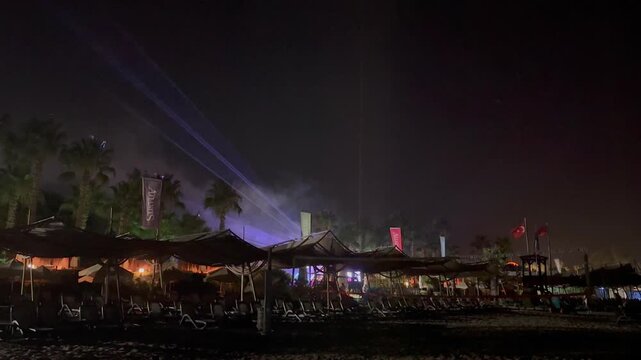 Turkey. 21.07.2026 Night beach party with laser show performance. Decorative lights beam crossing sky at seaside resort. Tropical palm tree under glowing illumination at night time.