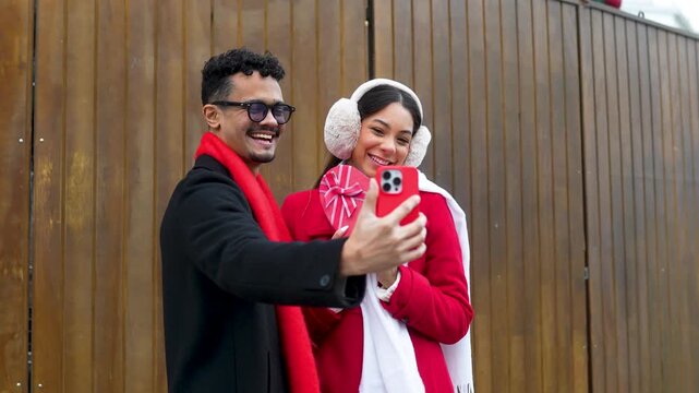 Young multicultural couple taking a selfie outdoors, holding a red heart-shaped valentine&rsquo;s day present
