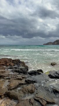 White sailing yacht resting off Cala Agulla Beach beneath dark storm clouds. Mallorca. Vertical Mediterranean seascape with turquoise water, waves and scenic mountain coastline.