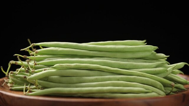Pile of raw needle bean or baby french beans on a brown wooden plate slowly rotating on a black background.